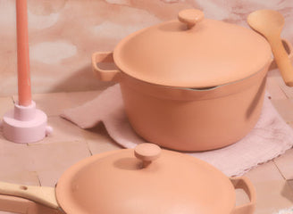 Two peach-colored cookware pieces, a lidded pot and a lidded pan with wooden handle, are displayed on a tiled surface with a candle, wooden spoon, and folded cloths.