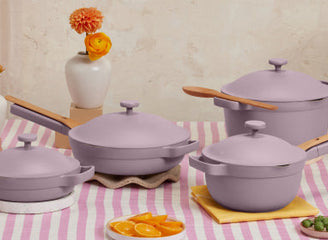 A set of four lavender-colored ceramic cookware pieces with wooden handles is displayed on a pink-and-white striped tablecloth, alongside orange slices and small vases with flowers.