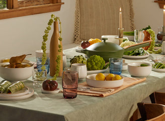 A dining table set with plates of vegetables, fruits, various dishes, glassware, and candles in a cozy, sunlit room with a window and cushioned bench.