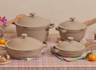 A set of four beige cookware pots and pans with lids arranged on a plaid tablecloth, surrounded by fruits and flowers.