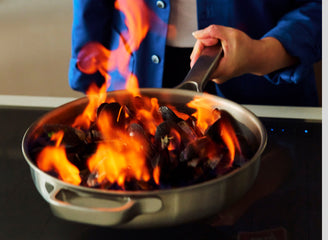 Person cooking with a flaming pan on a stove, wearing a blue jacket.