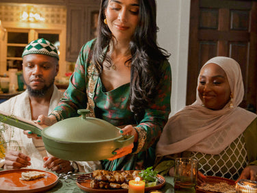 A woman in traditional clothing serves food from a green pan at a dining table, seated with two others dressed in festive attire.