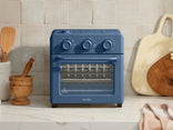 Blue toaster oven on a kitchen counter with wooden utensils and a cutting board in the background.