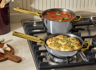 Two pots on a stove with one containing tomato sauce and basil, the other with bread and herbs.