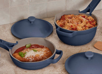 Two blue nonstick pans, one with breaded chicken in tomato sauce and the other with spaghetti in red sauce, sit on a kitchen counter against a tiled backsplash.