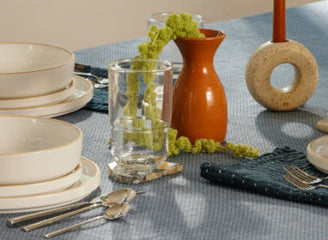 A dining table set for four with white dishes, glassware, silverware, a blue tablecloth, and decorative items including a brown vase, orange candle, and green foliage.