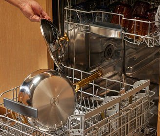 Person loading a titanium pan into an open dishwasher with wooden cabinets in the background