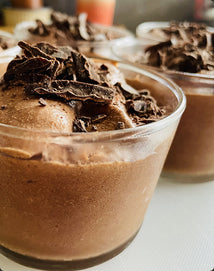 Chocolate mousse in a clear container with chocolate shavings on a white surface.