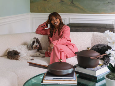A woman in a pink suit sits on a white sofa with a dog, next to a table holding two pans on top of stacked books.
