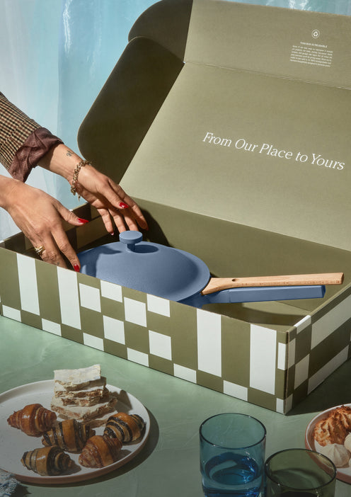 Person opening a box with kitchenware inside, set against a blurred background of a table with food.
