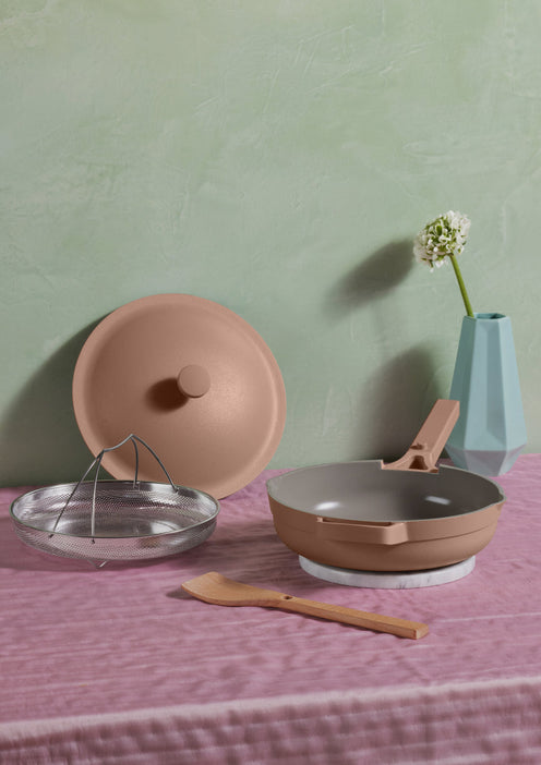 A beige nonstick pan with lid, mesh insert, and wooden spatula is arranged on a pink cloth next to a blue vase with a single white flower.