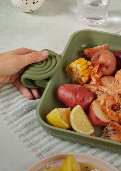 A hand uses a green silicone oven mitt to hold a baking dish filled with cooked shrimp, corn, potatoes, and lemon wedges on a striped cloth.