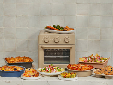 A beige countertop oven is surrounded by various dishes, including baked goods, salads, a casserole, nachos, and a plate of roasted vegetables, all set against a tiled backsplash.