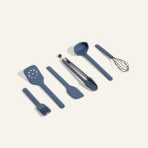 A set of six blue kitchen utensils, including a pastry brush, slotted spatula, spatula, tongs, ladle, and whisk, arranged in a row on a light background.