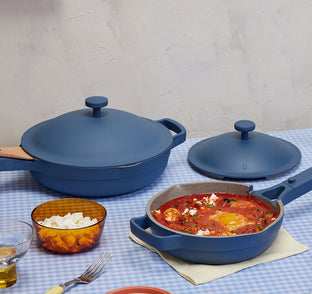 A set of matte blue cookware including a large lidded casserole dish, a smaller lidded pot, and a frying pan filled with shakshuka, styled on a blue-and-white checkered tablecloth with food props.