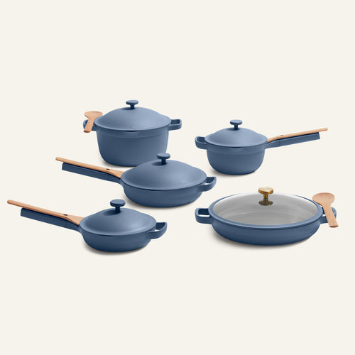 The Cookware Set + Dual Handle Always Pan, in blue with wooden handles and lids, is arranged on a light background with two wooden spoons resting on two pots.