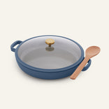 The Ceramic Nonstick Dual Handle Always Pan 32 cm in blue, with a glass lid, sits next to a wooden spoon on a simple, light background.