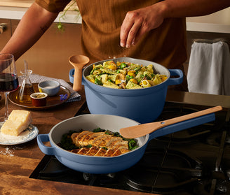 A person seasoning pasta in a large blue Dutch oven while cooking chicken and greens in a matching blue skillet on a stovetop. The kitchen scene includes a wine glass, cheese, and small dishes on a wooden table.