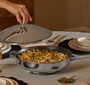 Person uncovering a pan of pasta with meat on a dining table.
