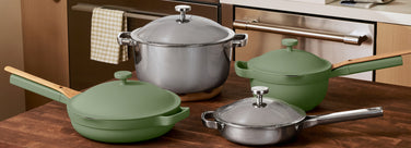 A set of green and stainless steel cookware, including a large pot with a lid, a frying pan, a skillet, and a smaller saucepan, arranged on a wooden kitchen countertop.