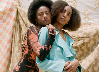 Two women stand closely together in front of patterned fabric backdrops. One wears a black and orange dress, and the other wears a light blue outfit.
