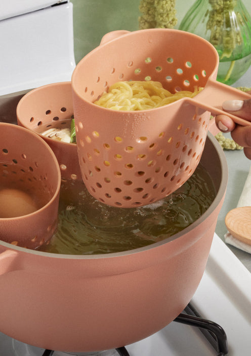A hand lifts a pink strainer scoop filled with cooked noodles out of a pot of boiling water on a stove.