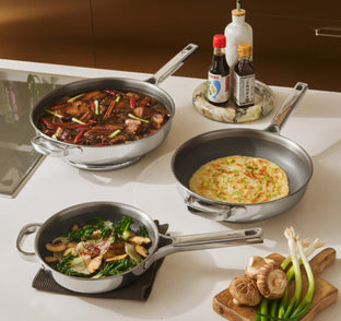 Three stainless steel pans on a kitchen counter, each containing different dishes; nearby are soy sauces, green onions, and mushrooms on a cutting board.