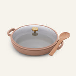 The Ceramic Nonstick Dual Handle Always Pan 32 cm is round, beige, with a glass lid, gold knob, two handles, and a wooden spoon on one handle against a plain background.