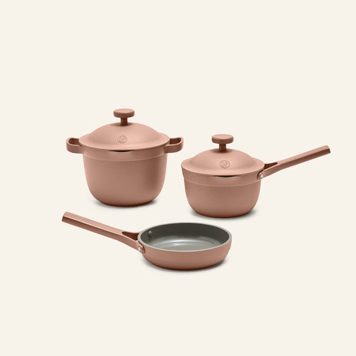 The Tiny Cookware Duo + Petite Perfect Pot, featuring rose gold-colored pots with lids and a frying pan, is displayed on a neutral background.