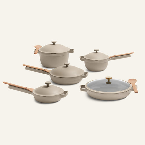 The Cookware Set + Dual Handle Always Pan features five beige ceramic nonstick pieces with wooden handles, including pots, pans, lids, and a wooden spoon.