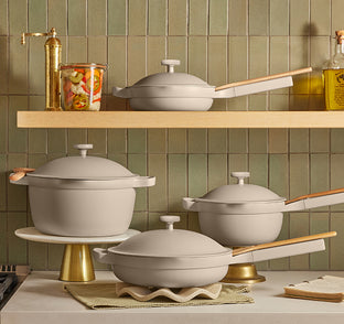 A coordinated cookware set in a styled kitchen scene with green tiled walls and wooden shelves. The set includes multiple light warm gray or off-white pots and pans with matching lids and light wood handles, displayed on a countertop and shelf.