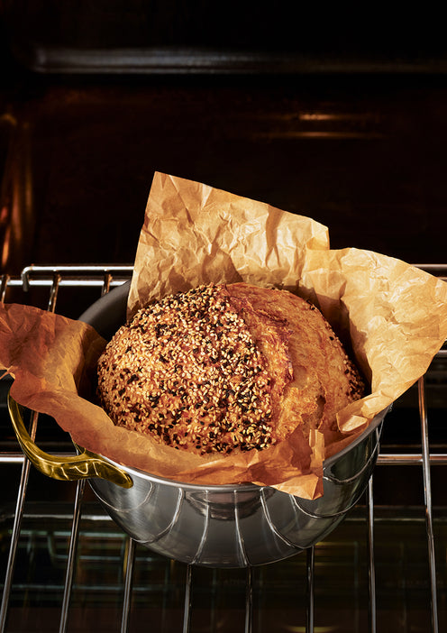 A round seeded bread loaf sits in a metal baking pan lined with brown parchment paper, placed on an oven rack inside an oven.