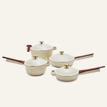 Four cream-colored pans from the Cookware Set, featuring wooden handles and gold knobs, are arranged on a light background.