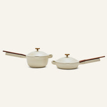 The Mini Cookware Duo features two cream-colored pots with wooden handles and lids, including the versatile Mini Always Pan, displayed side by side to showcase stylish and multifunctional cookware.