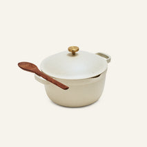 The Perfect Pot, cream-colored and family-sized with a lid and gold knob, features a wooden spoon resting through its side handle and sits on a plain light background.